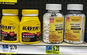 Four plastic bottles of medication on another drugstore shelf above their price tags. The two on the left are yellow with the word "Bayer" prominent in black type; above small type describes the product as "genuine aspirin". On the left are two clear plastic bottles with the Rite Aid drugstore chain logo on their yellow labels, which describe the product as "pain relief aspirin".