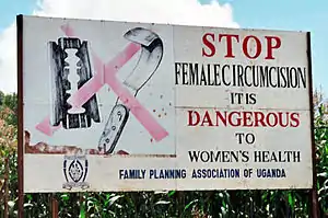 Billboard with surgical tools covered by a red X. Sign reads: STOP FEMALE CIRCUMCISION. IT IS DANGEROUS TO WOMEN'S HEALTH. FAMILY PLANNING ASSOCIATION OF UGANDA