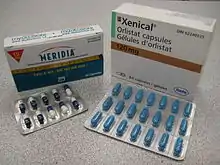 The cardboard packaging of two medications used to treat obesity. Orlistat is shown understanding under the brand name Xenical in a white package with the Roche logo in the bottom right corner (the Roche name within a hexagon). Sibutramine is below under the brand name Meridia. The package is white on the top and blue on the bottom separated by a measuring tape. The A of the Abbott Laboratories logo is on the bottom half of the package.